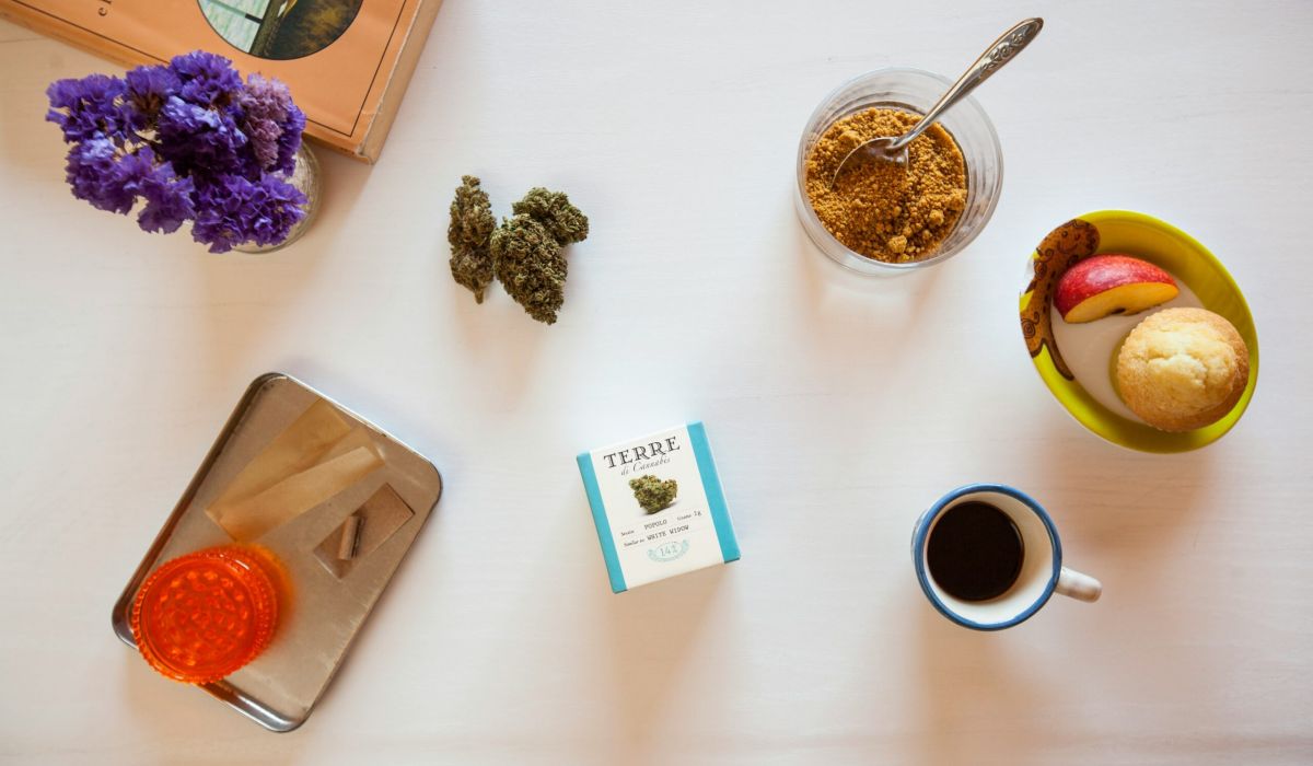 A spread of cannabis buds, espresso, brown sugar, an apple, a muffin, an orange grinder and rolling papers, and purple flowers